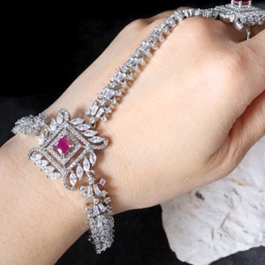 Puede incluir: Un conjunto de pulsera de cadena de mano y anillo de plata. La pulsera presenta una piedra central cuadrada de color rubí rodeada de cristales claros y brillantes. El anillo también tiene una piedra de color rubí. La joyería está sobre un fondo oscuro.
