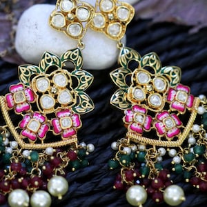 Pode incluir: Brincos dourados com detalhes florais e de contas. Os brincos apresentam uma parte superior em forma de flor com pedras transparentes, esmalte verde e detalhes de flores rosa. Contas vermelhas, verdes e brancas pendem da parte inferior.