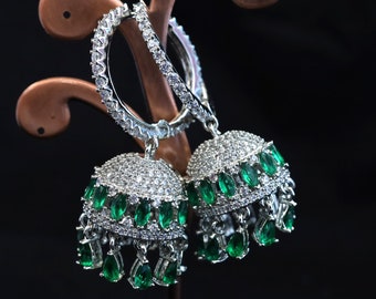 May include: A pair of silver-toned hoop earrings with a dangling bell-shaped design. The earrings are encrusted with small, clear crystals and feature teardrop-shaped green gemstones. The earrings are displayed against a dark background.