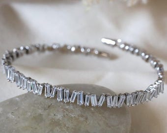 Op de afbeelding: Een zilverkleurige armband met een rij rechthoekige, heldere kristallen. De kristallen zijn dicht op elkaar geplaatst, waardoor een continu schittering ontstaat. De armband is open en rust op een lichtgekleurd, getextureerd oppervlak. De sieraden zijn elegant en geschikt voor speciale gelegenheden.
