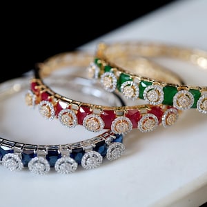 May include: Three gold bangles with jeweled accents. The top bangle has green stones, the middle bangle has red stones, and the bottom bangle has blue stones.