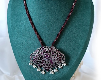 May include: A silver-toned pendant necklace with a large, ornate pendant. The pendant features a dark red gemstone design and is adorned with small white pearl-like accents. The necklace has a dark red beaded chain, displayed on a green mannequin.