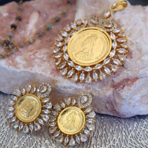 May include: A gold necklace with a pendant featuring a gold coin surrounded by clear stones. The pendant is on a rock with two matching earrings.