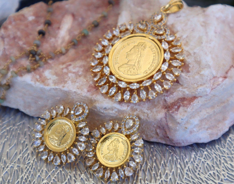 May include: A gold necklace with a pendant featuring a gold coin surrounded by clear stones. The pendant is on a rock with two matching earrings.