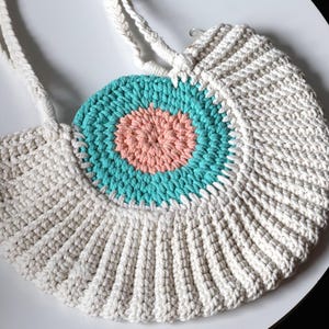 Puede incluir: Un bolso de crochet blanco con un diseño circular turquesa y melocotón. El bolso tiene dos correas largas.