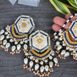 Pode incluir: Um par de brincos hexagonais brancos e azuis com detalhes dourados e de pérolas e contas pretas.