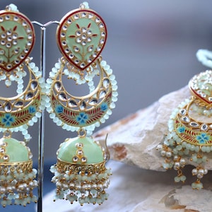 Pode incluir: Um par de brincos verdes e dourados com detalhes em pérolas e contas. Os brincos têm uma parte superior em forma de lágrima com um design de lua crescente e um charme de sino pendurado.