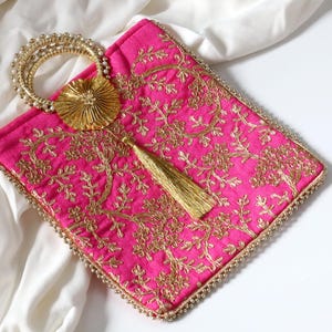 May include: A pink embroidered clutch bag with a gold tassel and a beaded handle. The bag features a floral design and is trimmed with gold.