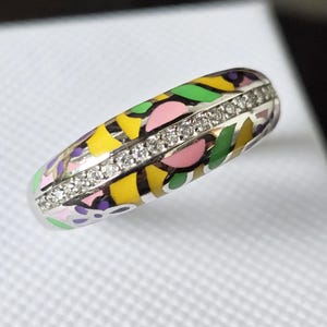 Puede incluir: Un anillo plateado con un diseño de esmalte colorido con formas amarillas, rosas, verdes y moradas. El anillo está acentuado con una fila de pequeñas piedras preciosas transparentes. El anillo está sobre una superficie blanca.