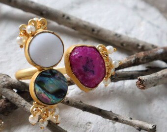 Könnte beinhalten: Goldring mit drei Steinen: einem weißen runden Stein, einem rosafarbenen unregelmäßigen Stein und einem grün-blauen schillernden Stein. Der Ring ist mit kleinen weißen Perlen verziert.