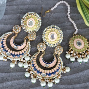 Pode incluir: Um conjunto de joias ornamentado com brincos e uma tiara. Os brincos têm formato de meia-lua com esmalte rosa e azul, adornados com pérolas e pedras transparentes. A tiara tem um design circular com enfeites semelhantes.