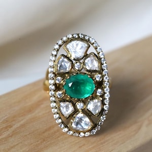 May include: A gold ring with an oval design, featuring a central emerald-cut green gemstone. The ring is embellished with clear accent stones and a halo of smaller gems, creating a sparkling effect.