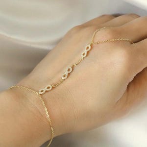 Puede incluir: Una cadena de mano dorada con tres s&iacute;mbolos de infinito adornados con peque&ntilde;as piedras brillantes. La cadena se conecta a un anillo y se envuelve alrededor de la mano, creando un aspecto delicado y elegante. La joya se exhibe sobre una tela blanca suave.