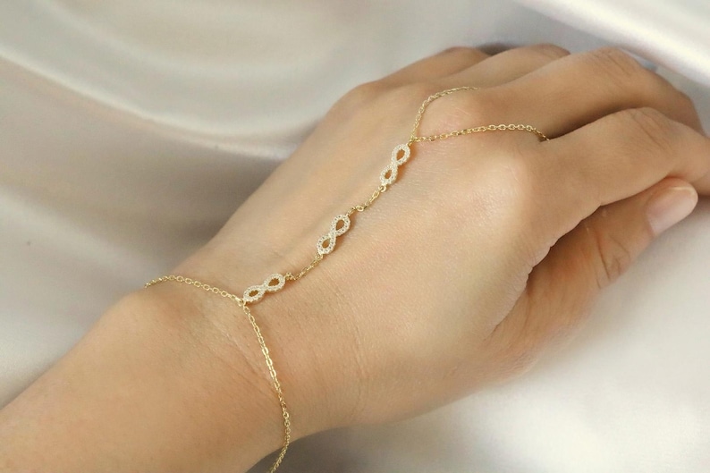 Puede incluir: Una cadena de mano dorada con tres s&iacute;mbolos de infinito adornados con peque&ntilde;as piedras brillantes. La cadena se conecta a un anillo y se envuelve alrededor de la mano, creando un aspecto delicado y elegante. La joya se exhibe sobre una tela blanca suave.