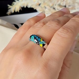 May include: A silver ring with a colourful design featuring blue, black, yellow, and white enamel. The ring has a rounded top with a pattern of dots and shapes. The ring is on a finger.
