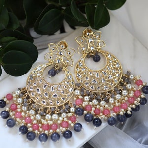 Pode incluir: Um par de brincos dourados com um design de lua crescente e um detalhe floral. Os brincos são adornados com pedras transparentes e cinzas e apresentam uma franja de contas multicoloridas.