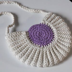 May include: A crocheted bag with a purple circle in the centre. The bag is made of cream-coloured yarn and has a long strap.