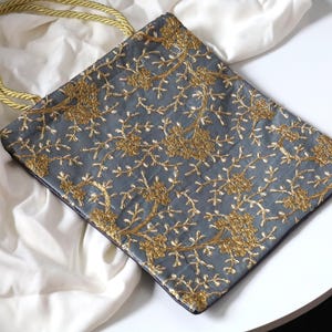 May include: A gray tote bag with a gold embroidered floral pattern. The bag has a gold rope handle.