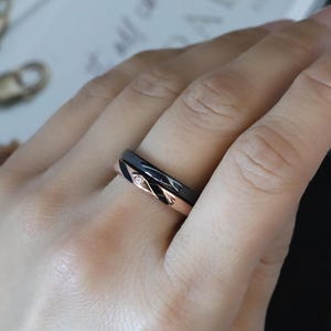 May include: A black and rose gold ring with a twisted design. The ring is worn on a person's finger.