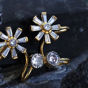 May include: A pair of gold earrings with a flower design featuring clear cubic zirconia stones.