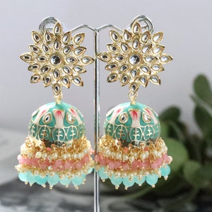 Pode incluir: Um par de brincos dourados ornamentados com um design floral. Os brincos apresentam um topo em forma de estrela com pedras transparentes e uma parte inferior em forma de sino turquesa e rosa adornada com contas. Os brincos são exibidos em um suporte prateado.