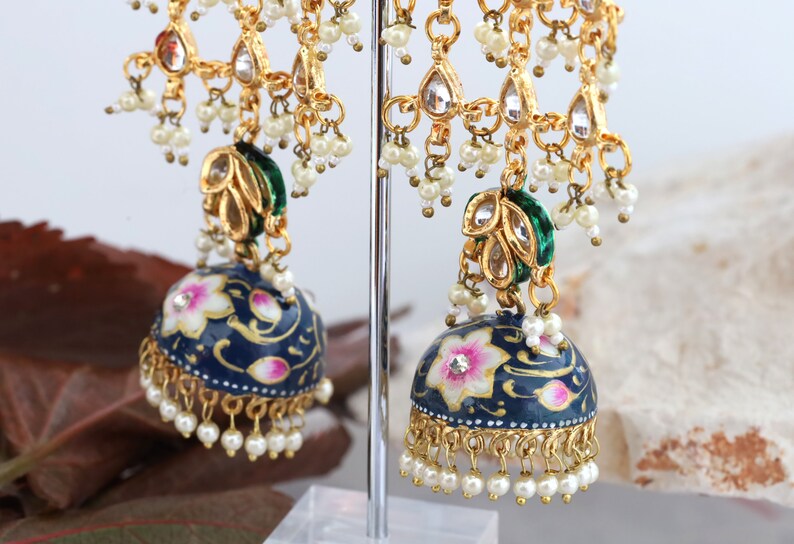May include: A pair of blue and gold earrings with white pearl accents. The earrings feature a floral design and a green and gold pendant.