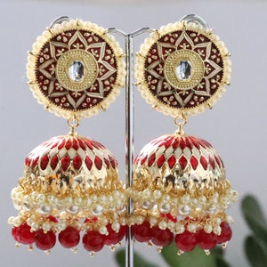 Pode incluir: Um par de brincos dourados com design em esmalte vermelho e branco. Apresentam uma parte superior circular com uma gema central e uma borda de pérolas, e uma parte inferior em forma de sino adornada com contas vermelhas e pérolas.
