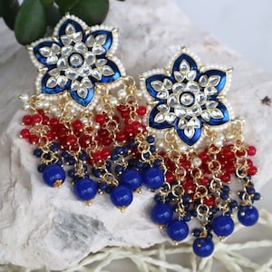 Pode incluir: Um par de brincos ornamentados com um design floral, esmalte azul e detalhes transparentes. Os brincos têm pingentes de contas vermelhas e azuis, com detalhes em metal dourado. Os brincos são para orelhas furadas.