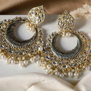 Pode incluir: Um par de brincos de ouro com um centro preto e um design em forma de lágrima. Os brincos são adornados com pequenas peças espelhadas e pérolas brancas.