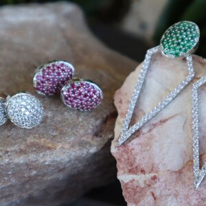 May include: Three pairs of earrings with different coloured gemstones. The first pair has round, pink gemstones. The second pair has round, white gemstones. The third pair has green gemstones set in a triangular shape.