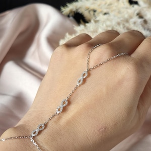 Puede incluir: Una delicada cadena de mano plateada con m&uacute;ltiples s&iacute;mbolos de infinito adornados con peque&ntilde;as piedras brillantes. La cadena se conecta a una pulsera y un anillo, creando un accesorio elegante. La joya se muestra sobre un fondo suave y neutro.
