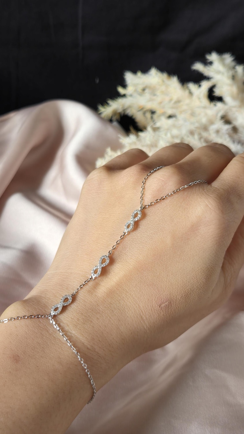 Puede incluir: Una delicada cadena de mano plateada con m&uacute;ltiples s&iacute;mbolos de infinito adornados con peque&ntilde;as piedras brillantes. La cadena se conecta a una pulsera y un anillo, creando un accesorio elegante. La joya se muestra sobre un fondo suave y neutro.