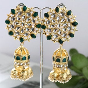 Pode incluir: Um par de brincos de ouro com pedras verdes e brancas. Os brincos têm uma parte superior redonda com um design floral e um charme em forma de sino que pendura.