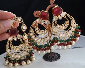 May include: A pair of gold earrings with red and green stones and pearls. The earrings are in the shape of a crescent moon and have a dangling design.