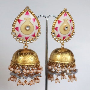 Pode incluir: Brincos de ouro com um pingente em forma de lágrima com um design branco e rosa. O pingente é rodeado por strass transparentes. Os brincos têm um pingente em forma de sino com um acabamento dourado e uma franja de contas brancas e cinzas.