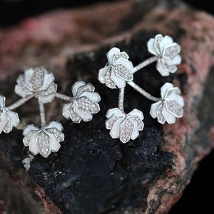 May include: A pair of elegant, silver-toned cufflinks with a floral design. Each cufflink features three white, petal-shaped elements adorned with sparkling, clear gemstones. The cufflinks are displayed on a dark, textured surface.
