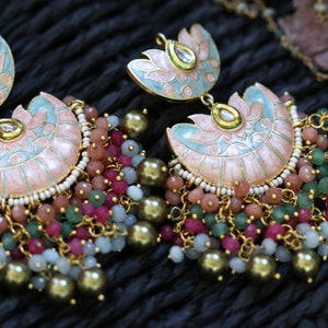 Pode incluir: Um par de brincos de ouro com desenhos de flores de lótus em esmalte rosa e azul. Os brincos têm várias camadas de contas em várias cores, incluindo rosa, verde e branco.