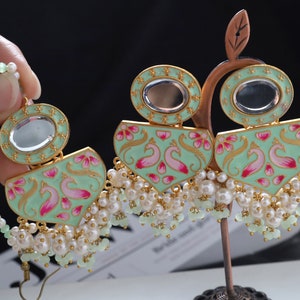 Pode incluir: Brincos verde menta e dourados com um design de pavão. Os brincos apresentam um topo oval espelhado e uma base em forma de leque com detalhes em rosa e dourado. Detalhes de pérolas brancas e contas verdes pendem da base.