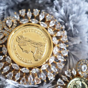 May include: A gold pendant featuring a Queen Victoria coin surrounded by a halo of clear, faceted stones. The coin has the inscription "BRITANNIA" and "VICTORIA D.G."