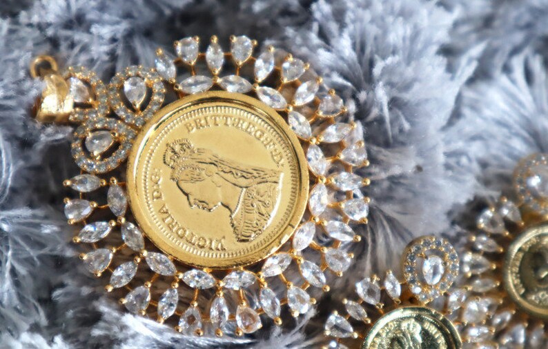 May include: A gold pendant featuring a Queen Victoria coin surrounded by a halo of clear, faceted stones. The coin has the inscription "BRITANNIA" and "VICTORIA D.G."