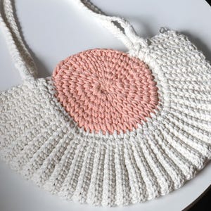 May include: A crocheted bag with a white and pink color scheme. The bag has a round shape and a single strap.
