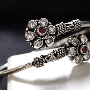 May include: Silver bangle bracelet featuring two flower-shaped designs. Each flower has a red gemstone at its centre and white gemstones surrounding it.