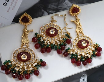 May include: A pair of gold earrings with red and green beads, a red stone, and a small figure of a person playing a musical instrument.