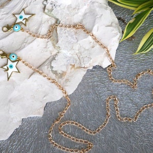 May include: A gold chain with two white star charms and blue evil eye charms. The chain is laying on a white rock with green leaves in the background.
