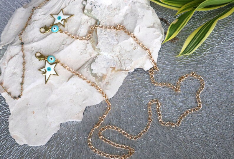May include: A gold chain with two white star charms and blue evil eye charms. The chain is laying on a white rock with green leaves in the background.