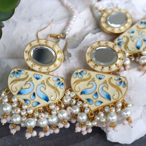 Pode incluir: Um par de brincos de ouro com base creme e desenhos de pavão azuis e dourados. Os brincos têm um espelho redondo no centro e uma franja de pérolas brancas.