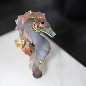 Puede incluir: Un anillo de tono dorado con un diseño de caballito de mar detallado hecho de concha de abulón iridiscente.