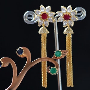 May include: A pair of gold dangle earrings with a flower design and red and white stones. The earrings have a gold chain tassel.