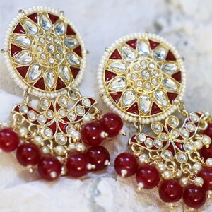 Pode incluir: Um par de brincos de ouro com detalhes em esmalte vermelho e branco e pérolas. Os brincos têm um design redondo floral com contas vermelhas penduradas.