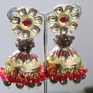 Pode incluir: Um par de brincos dourados em forma de sino com detalhes intrincados. Os brincos apresentam um design floral no topo com detalhes vermelhos e transparentes, um aglomerado de contas cinzentas e uma base adornada com contas vermelhas e rosa. Os brincos são exibidos em um suporte prateado.